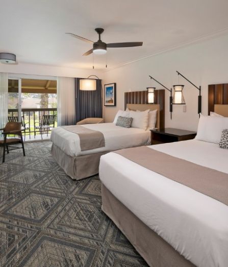 A spacious hotel room with two large beds, neutral tones, a balcony view, work desk, and a patterned carpet.