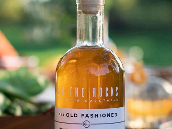 A bottle of &ldquo;The Rocks&rdquo; Old Fashioned cocktail on a table, with blurred greenery and glasses in background at an outdoor setting.