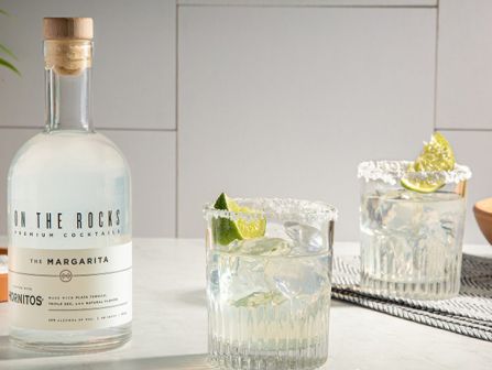 A bottle of tequila on a kitchen counter with two rocks glasses filled with ice and lime wedges, plus a bowl of limes in the background.