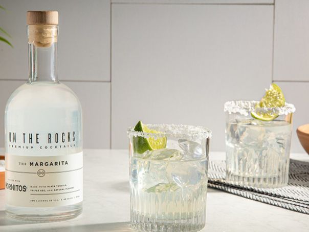 A bottle of tequila on a kitchen counter with two rocks glasses filled with ice and lime wedges, plus a bowl of limes in the background.