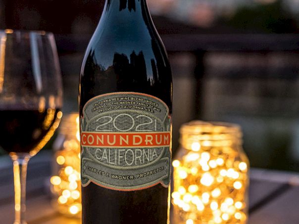 A bottle of wine labeled &ldquo;Conundrum&rdquo; sits on a table with a glass of red wine and glowing candles in the background.