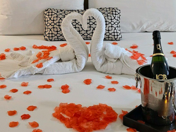 A romantic hotel setup with two towel swans facing each other, rose petals on the bed, a champagne bucket, glasses, and a heart-shaped arrangement.