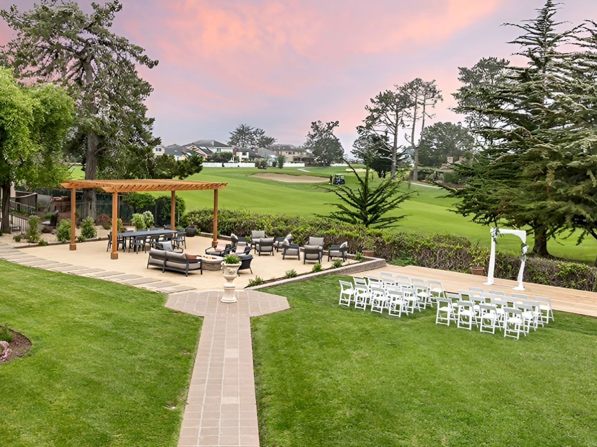Outdoor wedding setup at sunset: lawn, white chairs arranged in rows, an altar arch, and a wooden gazebo with cocktail tables on a paved path.