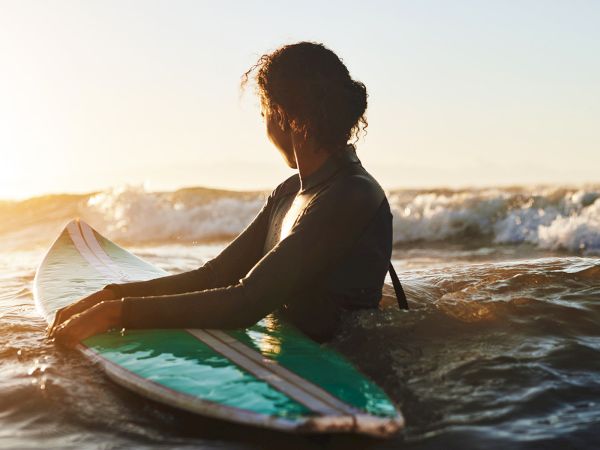 A person sits on a surfboard in the ocean at sunset, facing the waves, with a calm, reflective vibe as light glints off the water.