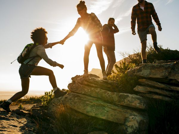 Four hikers helping each other climb rocky steps at sunset, teamwork and adventure on a scenic trail.